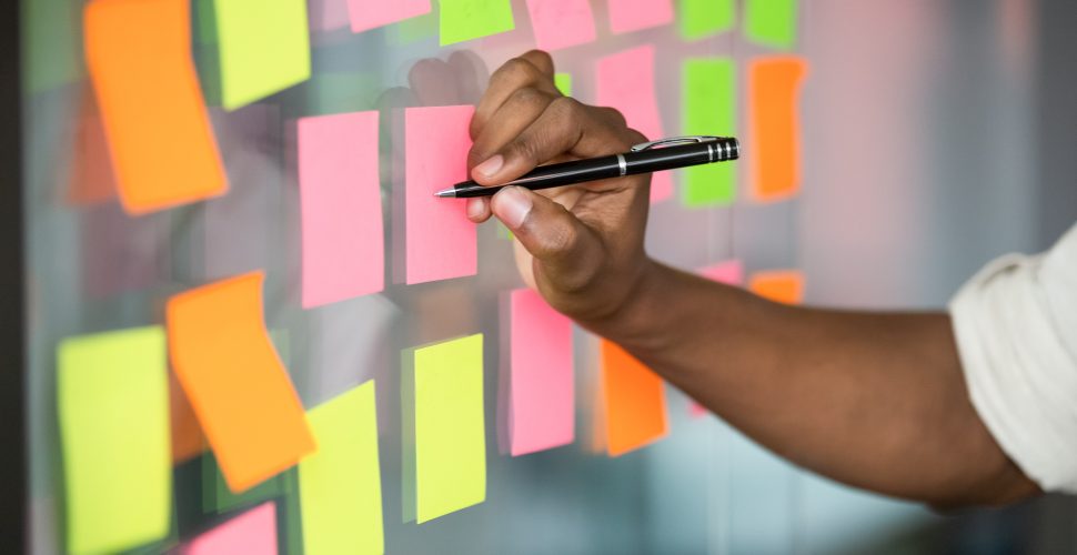 Close up african american manager writing notes on colorful sticky notes on kanban board. Mixed race employee managing project workflow, scheduling tasks or planning strategy on glass wall at office.