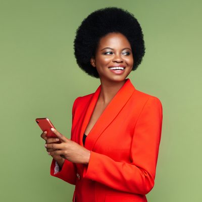 Successful african businesswoman hold smartphone look aside with confident happy smile. Afro american young entrepreneur female in luxury red suit with mobile phone isolated over green studio wall